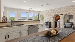 A modern kitchen with white cabinets, a stone wall, and a large window overlooking a scenic view. - Lake Travis Oasis, Pool, Hot Tub, Lake Views, Near Marinas & Austin! (Austin)