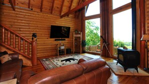 A log cabin interior with a wooden staircase, leather sofa, a fireplace, and a large window offering a view of trees. - Mountain top overlooking lake, Hot Tub, Pickleball, Yukon Fire Pit, 5 STAR, PETS (Waleska)
