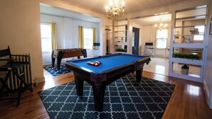A pool table in a room with a chandelier, wooden flooring, and a blue patterned rug.
