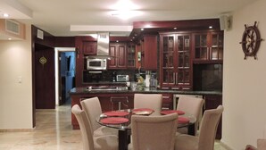 A modern kitchen with dark wood cabinets, a stainless steel refrigerator, a microwave, and a large island with a glass top. There are beige chairs around a small round table with red placemats. - New Oceanfront Dream apartment on the beach of Isla Verde! (Carolina)