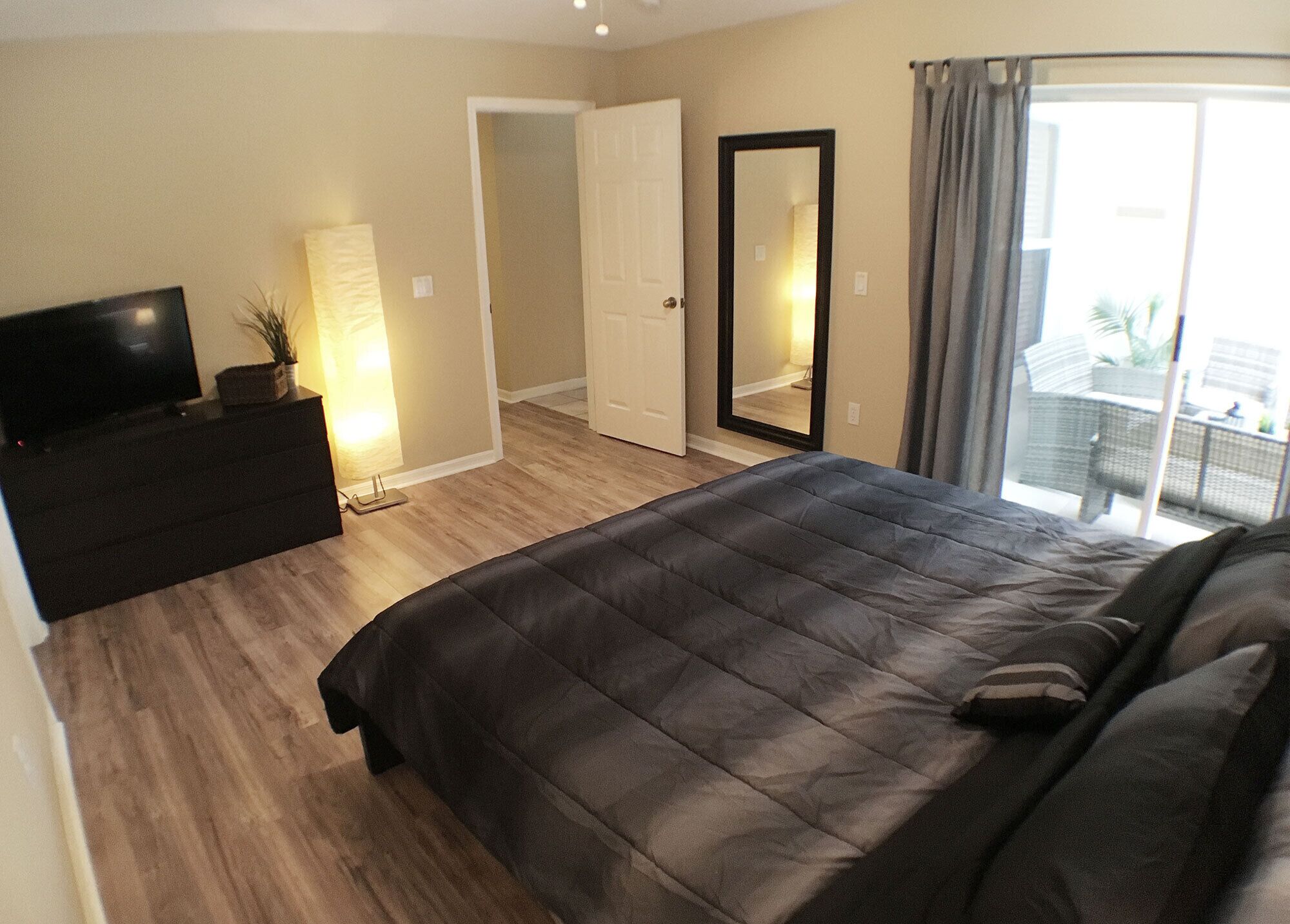 Quarto com uma cama grande, uma televisão sobre uma cômoda, um espelho e uma porta de vidro deslizante para uma varanda.