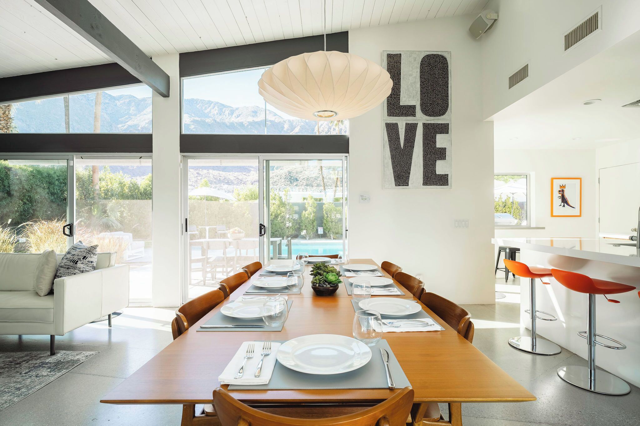 A modern dining area with a wooden table set for six, large windows offering mountain views, and a kitchen area with a white countertop and red bar stools.
