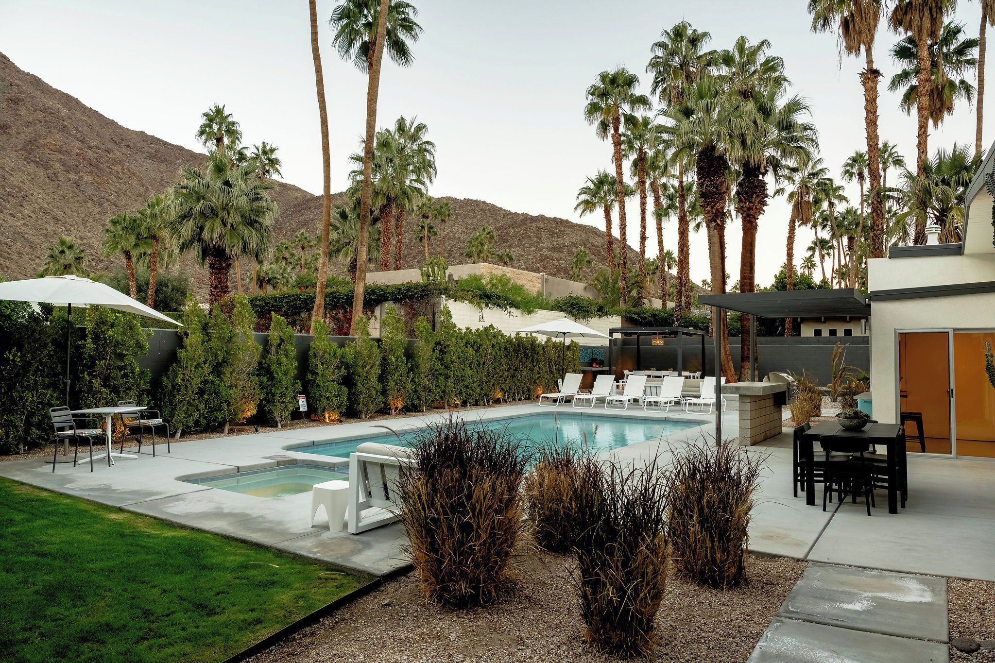 A pool area with lounge chairs, a table, and a covered seating area, surrounded by palm trees and a mountain backdrop.