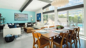 A modern dining area with a wooden table set for six, a white sofa, and a large window offering a mountain view. - Hollywood History Views Saltwater Pool & Spa (Palm Springs)