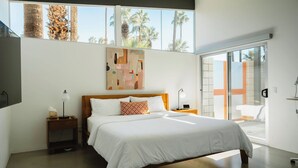 A modern bedroom with a large bed, a nightstand, a wall-mounted TV, and a sliding glass door leading to a balcony with a view of palm trees. - Hollywood History Views Saltwater Pool & Spa (Palm Springs)