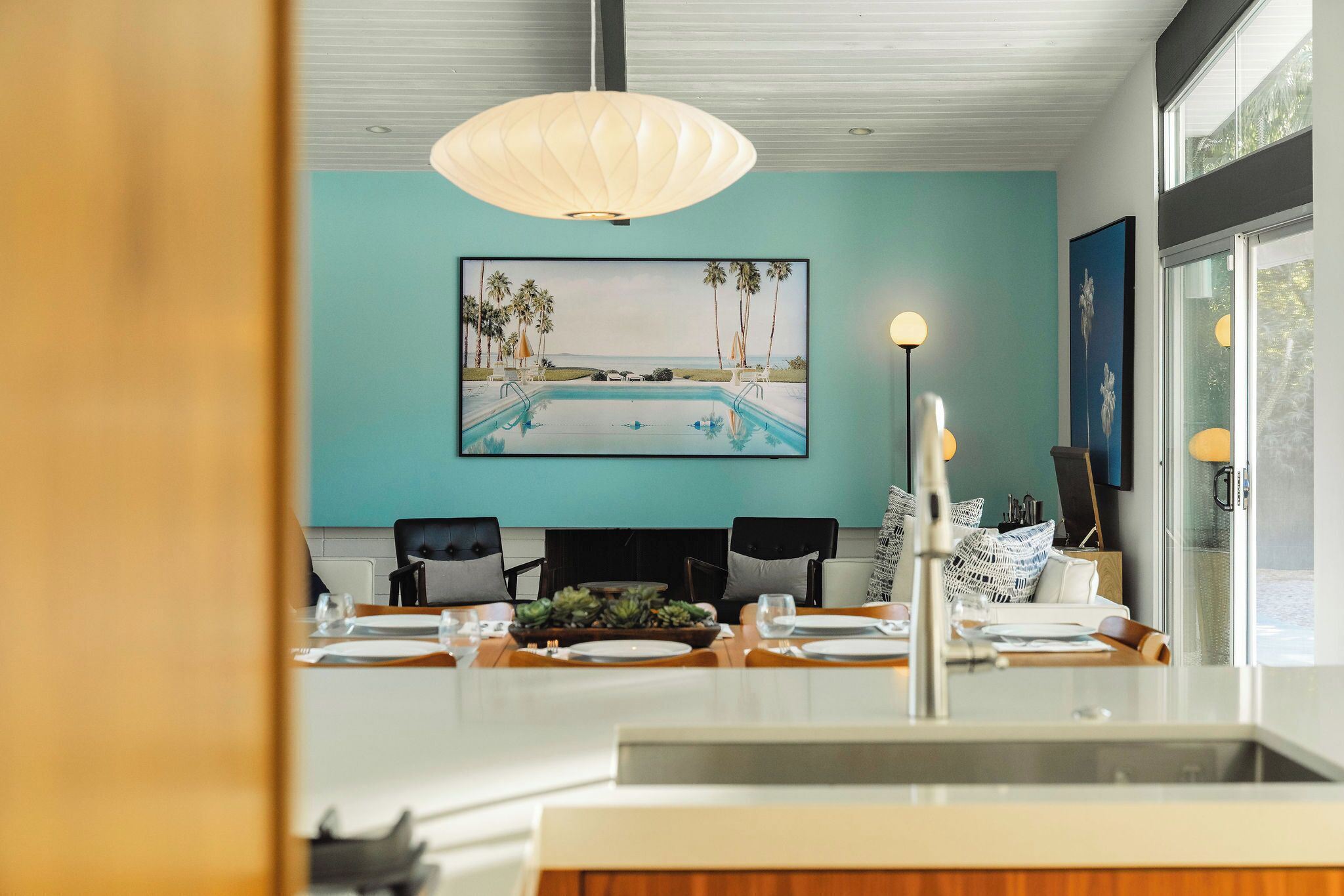 A modern kitchen with a teal accent wall, a large framed picture of a pool, and a dining area with a white island.