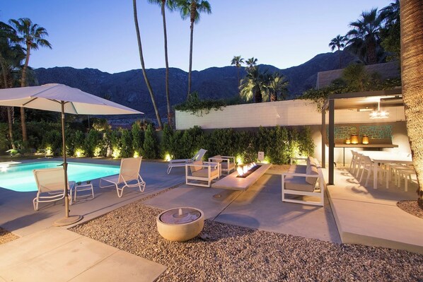 A poolside area with lounge chairs, a fire pit, and a bar area with ambient lighting.