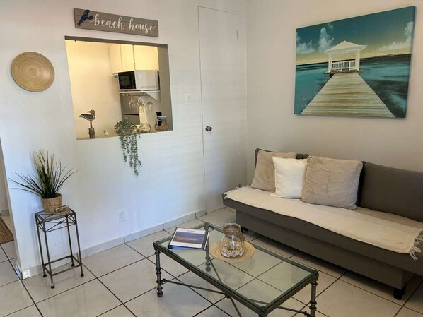 A living room with a sofa, a coffee table, a mirror, and a painting of a beach house. - Charming Apartment w/Pool. Just walking minutes from the Beach and Restaurants. (San Juan)