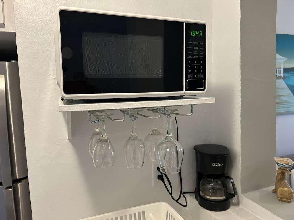 Una cocina con un microondas en un estante, copas de vino colgando debajo y una cafetera sobre la encimera.