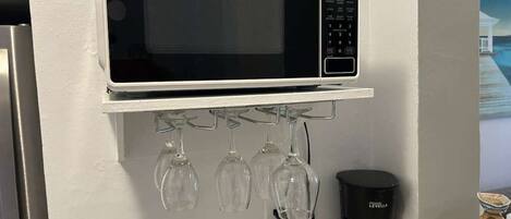 Una cocina con un microondas en un estante, copas de vino colgando debajo y una cafetera sobre la encimera.