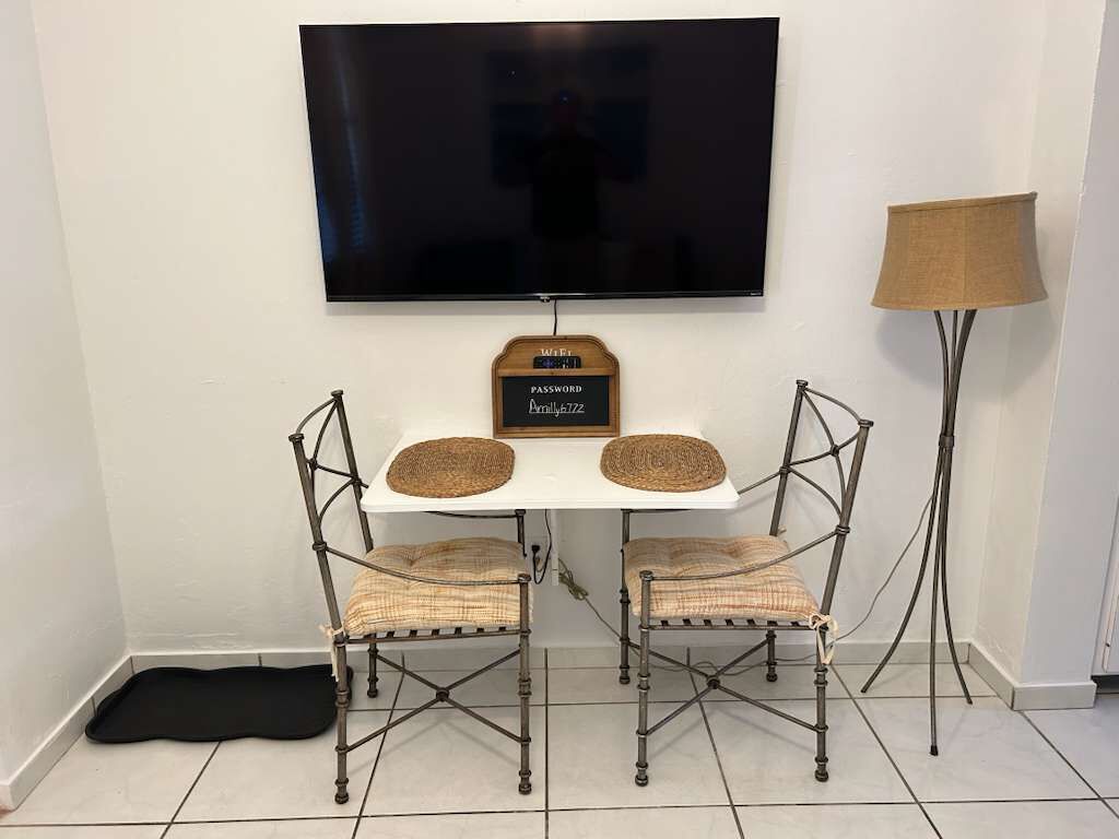 Un pequeño comedor con una mesa blanca, dos sillas de mimbre, un televisor montado en la pared y una lámpara de pie.