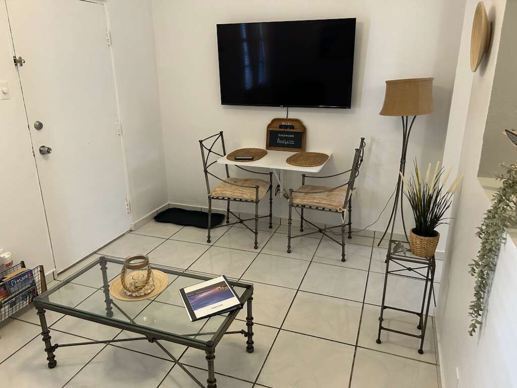 A living room with a TV mounted on the wall, a glass coffee table with a book, a small table with two chairs, and a floor lamp.