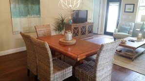 A dining area with a wooden table and wicker chairs, a television on a wooden cabinet, and a view of the beach through a window.