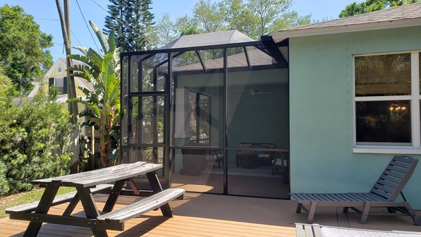 A covered patio with a wooden table and chairs, a glass enclosure, and a view of a house in the background.