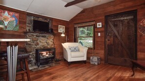A cozy cabin interior with a stone fireplace, a white armchair, and wooden floors. - Sweet Cabin in the Woods -10 min to Ashvl., Quiet, BEES , Garden & DECK. Dogs Ok (Asheville)