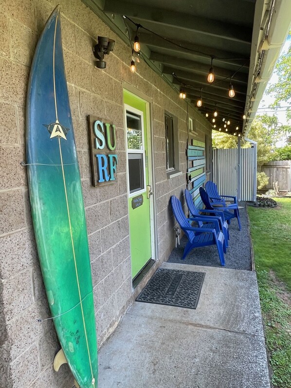 A building with a surfboard leaning against the wall, a door labeled "SURF", and blue chairs outside.
