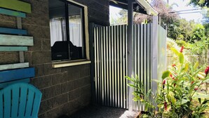 Un espace extérieur abrité comprenant une chaise en plastique bleue, une fenêtre avec un rideau, un portail en métal et un jardin avec différentes plantes.