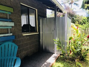 Un espacio al aire libre cubierto con una silla de plástico azul, una ventana con cortina, una reja metálica y un jardín con diversas plantas.