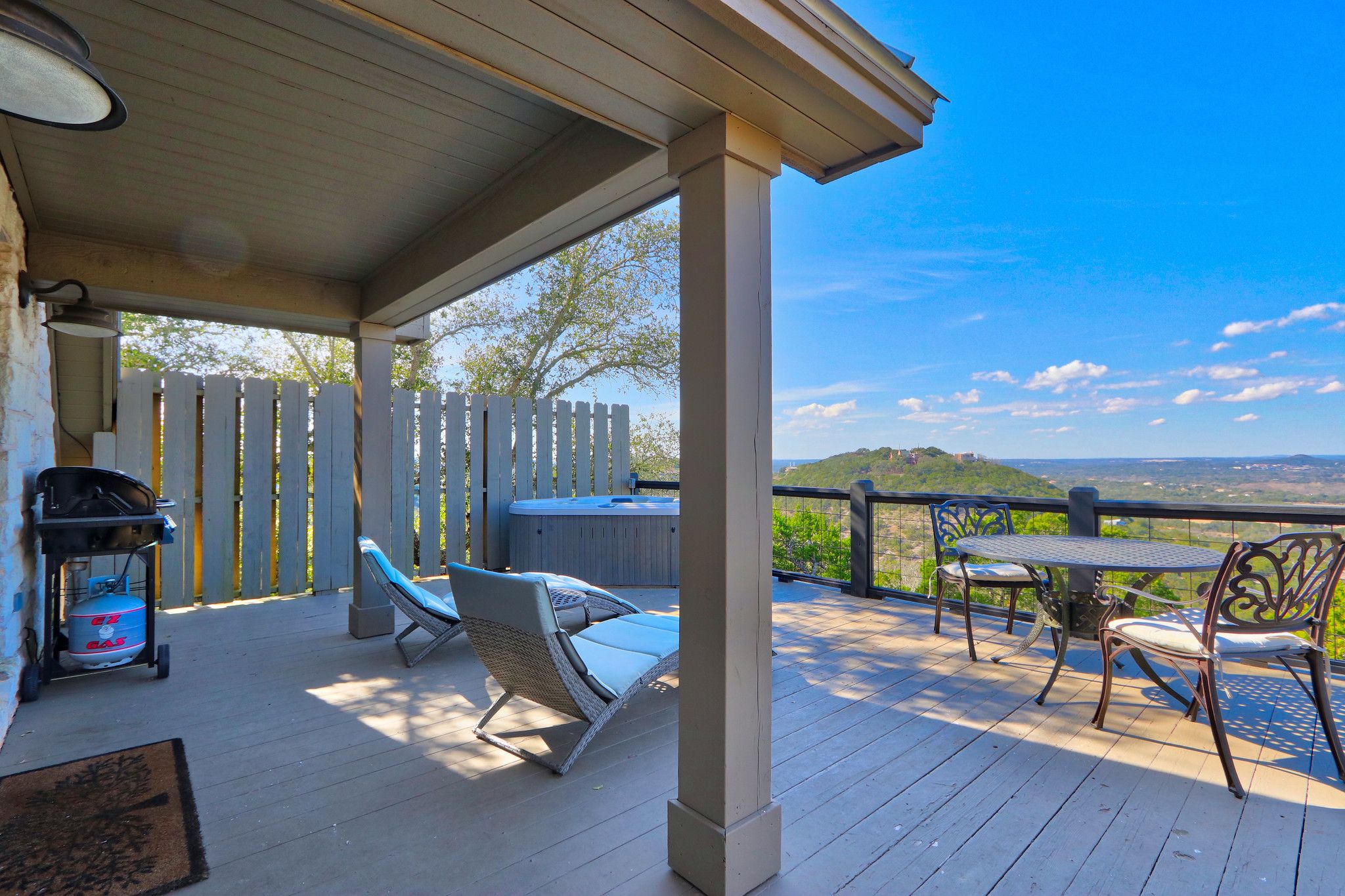 A covered outdoor deck with a hot tub, lounge chairs, and a table with chairs.