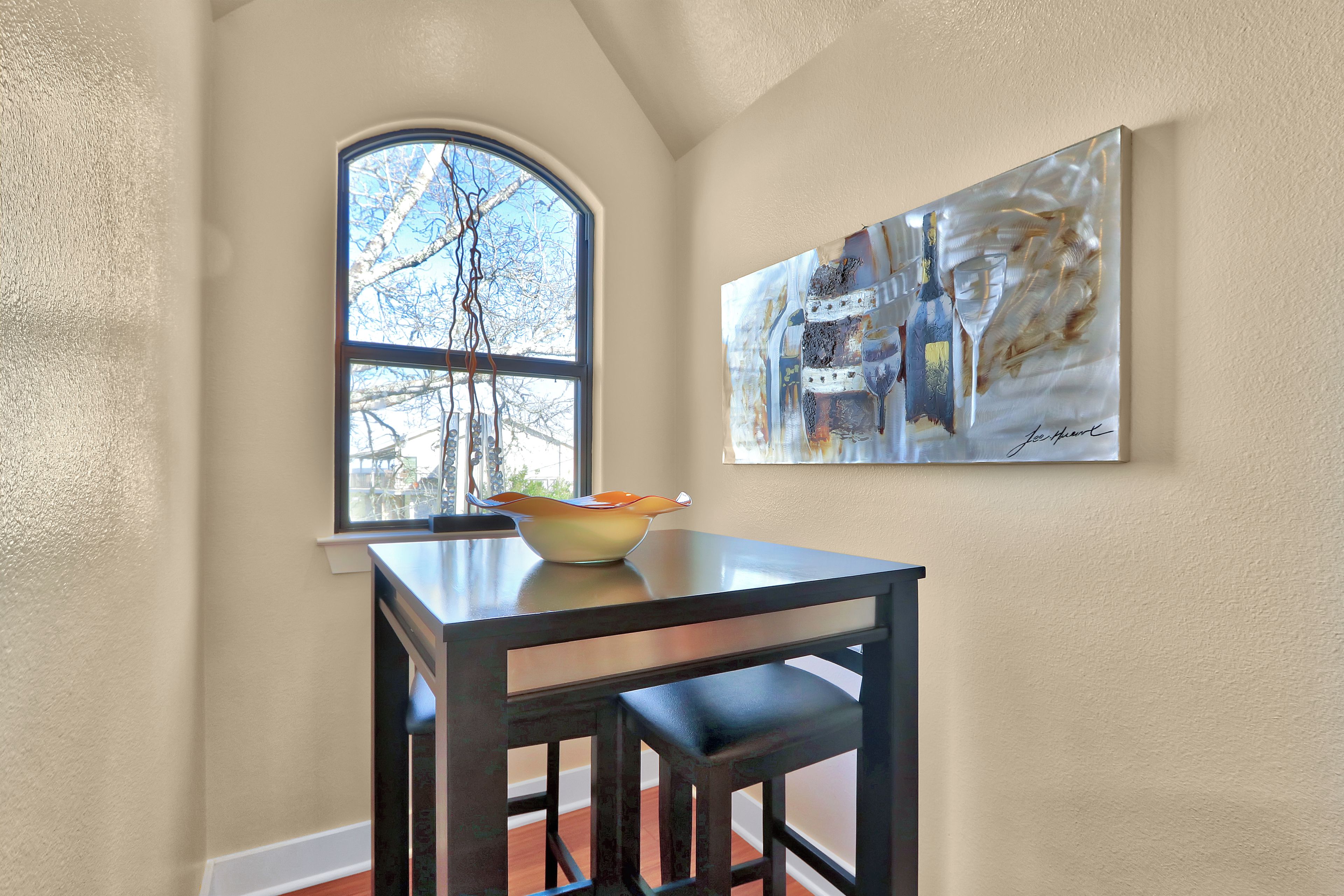 A dining area with a table and chairs, a bowl on the table, a window with a view of trees, and a painting on the wall.