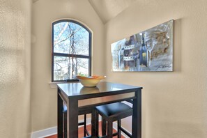 Uma área de refeições com uma mesa e cadeiras, uma tigela sobre a mesa, uma janela com vista para árvores e um quadro na parede.