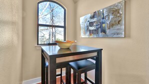 A dining area with a table and chairs, a bowl on the table, a window with a view of trees, and a painting on the wall. - Romantic-amazing Views-Newly Renovated-new Hot Tub-has It All! (Wimberley)