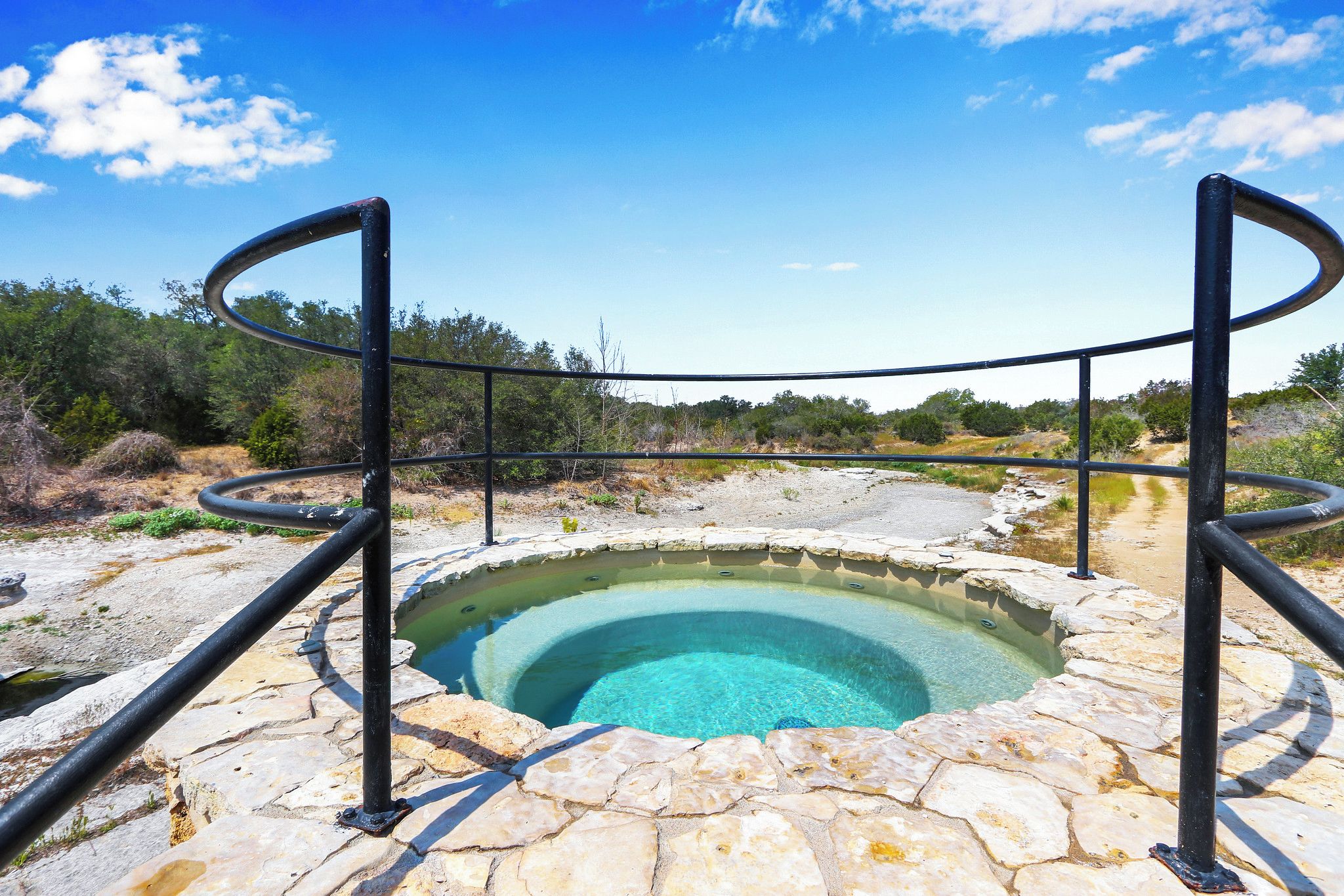 A circular hot spring with a stone edge and metal railings, surrounded by a natural landscape.