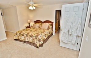 Quarto com cama, mesa de cabeceira, luminária, ventilador de teto e porta com dois roupões brancos pendurados.