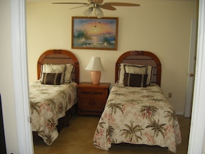 Une chambre avec deux lits, une table de chevet avec une lampe, un ventilateur de plafond et un tableau représentant un coucher de soleil.