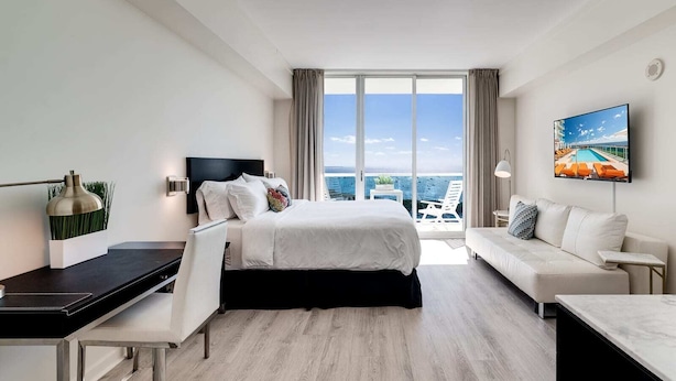 Une chambre d’hôtel moderne, dotée d’un grand lit, d’un bureau, d’un canapé et d’une télévision. La chambre dispose d’une grande fenêtre offrant une vue sur l’océan.