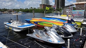 Un port de plaisance avec des bateaux, des jet-skis et un bâtiment portant une enseigne indiquant la location de kayaks et de planches de stand-up paddle.