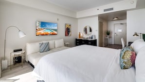 Une chambre d’hôtel moderne, dotée d’un grand lit, d’un bureau, d’une chaise, d’une lampe sur pied, d’une télévision et d’un tableau représentant une plage.