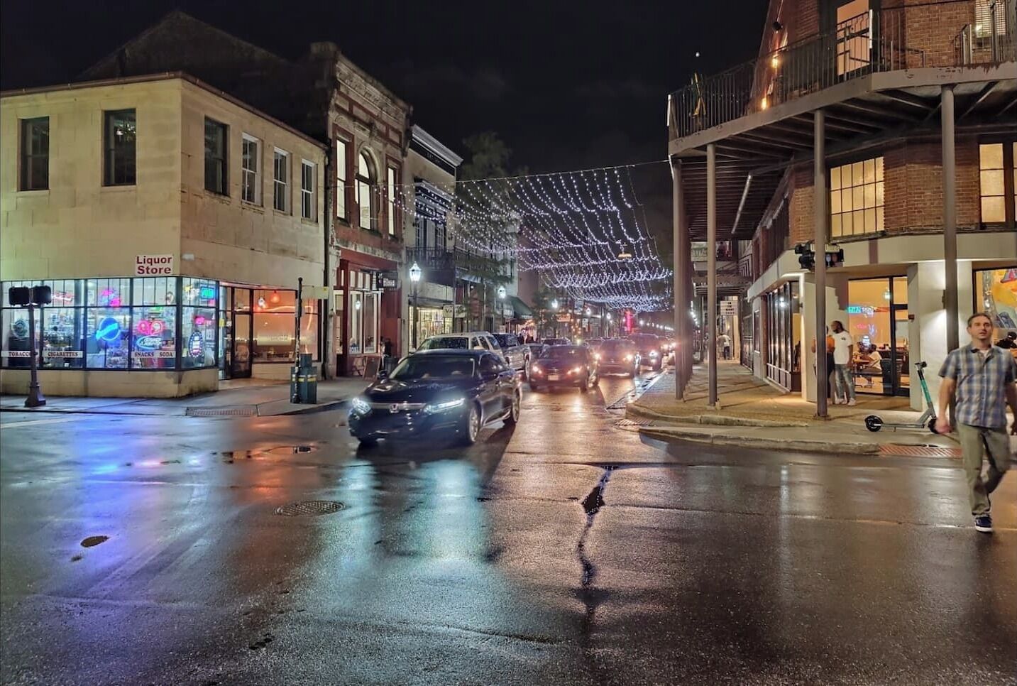 A street at night with wet pavement, cars, and illuminated storefronts.