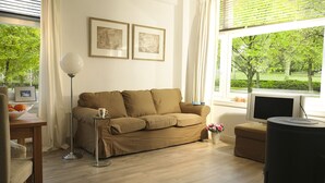 A living room with a beige sofa, a glass coffee table, a floor lamp, a TV, and a window with a view of trees. - Private Apartment in Good Area (Amsterdam)