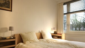 A neatly made bed in a room with a window, a bedside table with a lamp, and a bookshelf. - Private Apartment in Good Area (Amsterdam)
