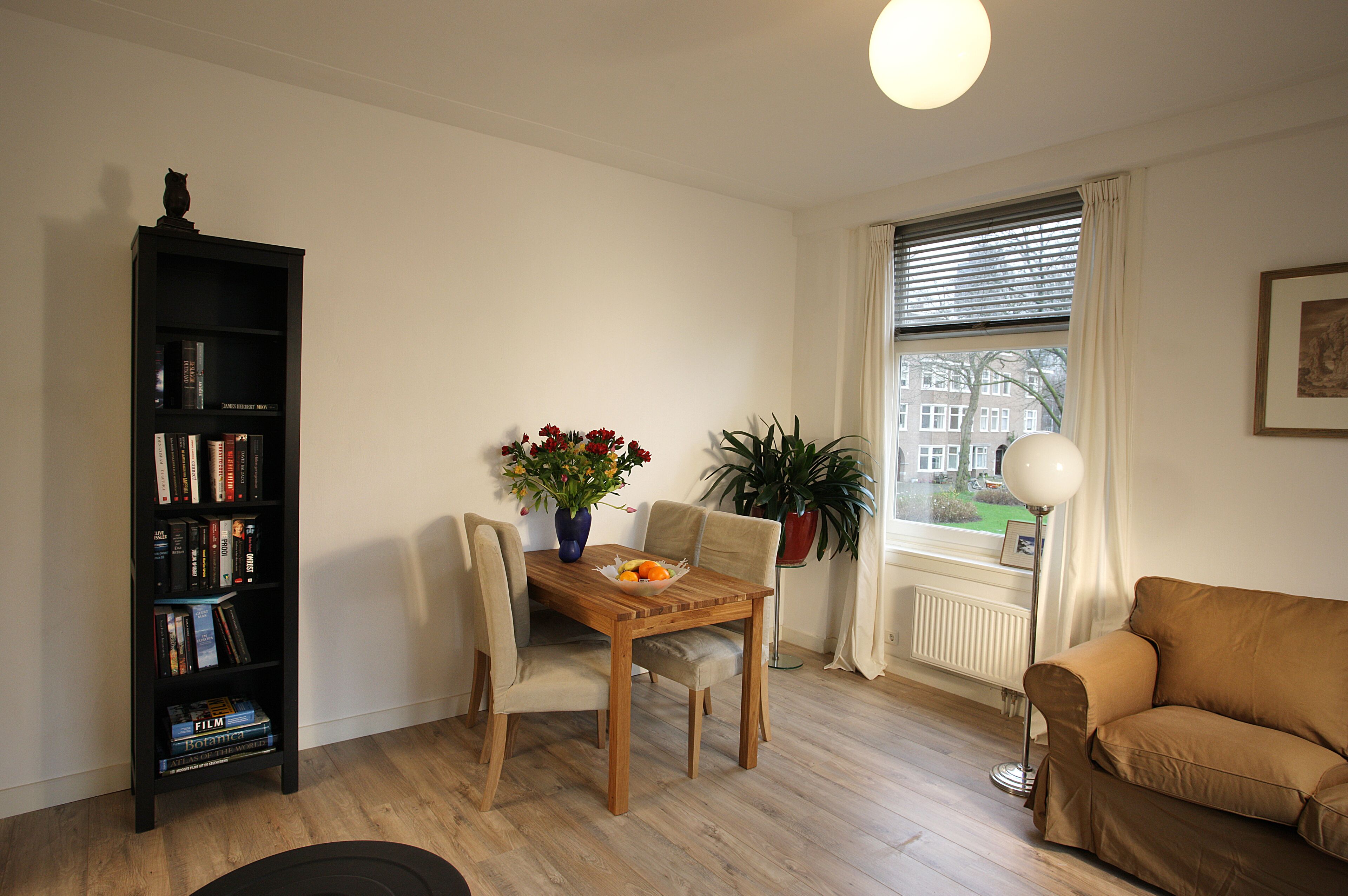 A living room with a wooden dining table, two chairs, a bookshelf, a sofa, a floor lamp, and a window with curtains.
