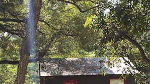 Un petit bâtiment à deux étages, avec une porte et des fenêtres rouges, entouré d’arbres et d’une zone herbeuse.