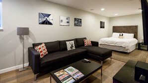 Una sala de estar moderna con un sofá modular negro, una mesa de centro con revistas, una cama con cabecera blanca y tres obras de arte enmarcadas en la pared.
