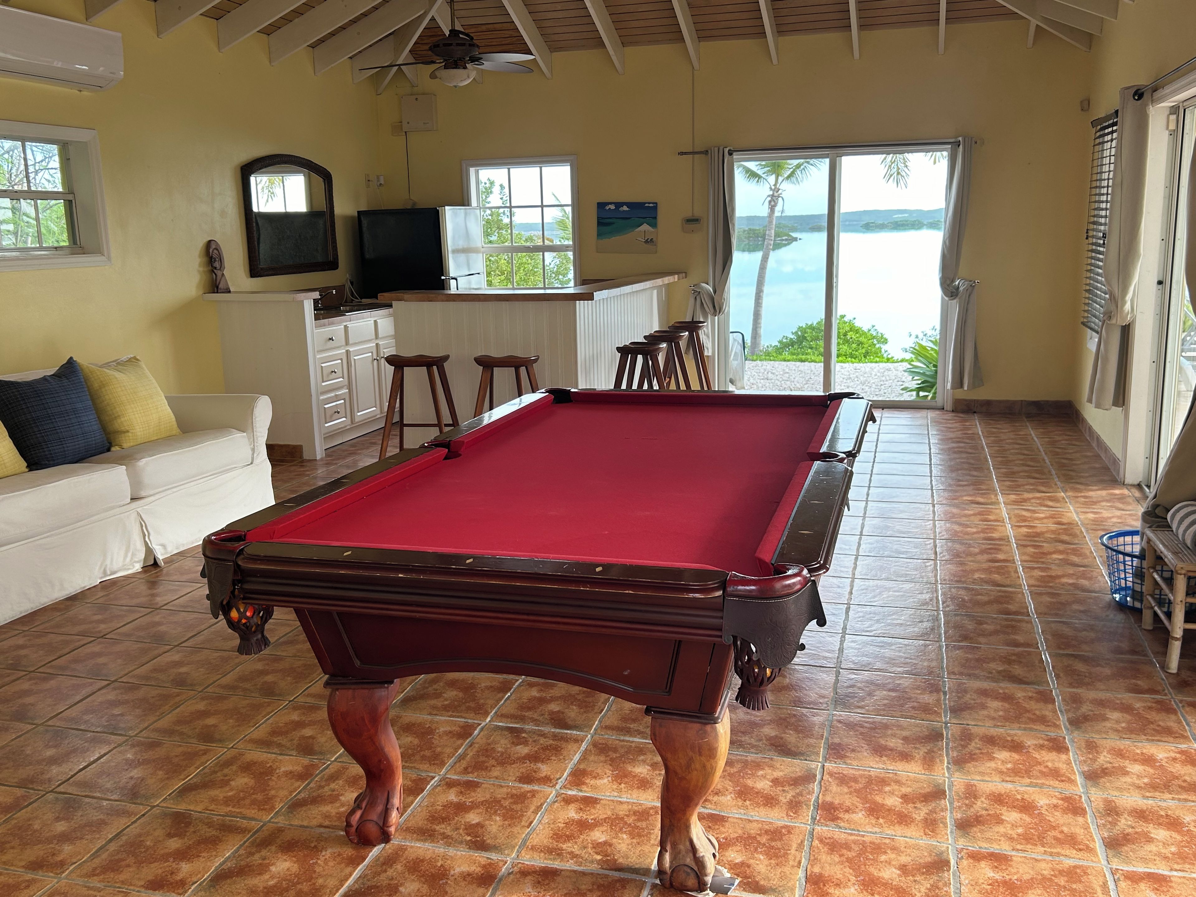 A pool table in a room with a tiled floor, a white sofa, a television, and a view of the ocean through a glass door.