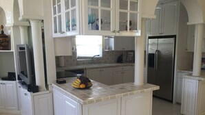 Eine Küche mit weißen Schränken, einer zentralen Kochinsel mit Obst, einem Edelstahl-Kühlschrank und einem Fenster mit Jalousien.