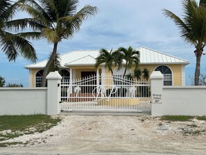 A gated property with a white metal fence, a palm tree, and a building with a yellow facade. - Luxury Turks & Caicos Villa with Infinity Pool on Chalk Sound (Providenciales)
