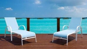 Zwei weiße Lounge-Stühle auf einer Holzterrasse mit Blick auf türkisfarbenes Wasser und entfernte Inseln.