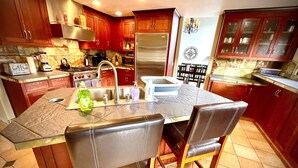 Une cuisine avec des meubles de rangement en bois, un réfrigérateur en acier inoxydable, une cuisinière à gaz et un évier. Il y a deux chaises autour d’une table, et le mur derrière le plan de travail est recouvert de carreaux.