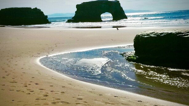 Une plage avec une formation rocheuse naturelle visible au loin, un rivage sablonneux et des empreintes de pas qui mènent à l’eau.