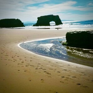 Una playa con una formación rocosa natural en la distancia, una orilla de arena e huellas que se adentran en el agua.