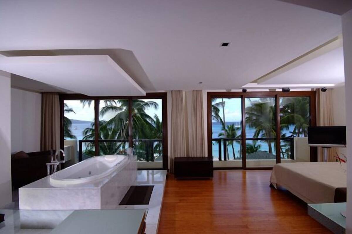 A modern hotel room with a large bathtub, a bed, a TV, and a balcony with a view of palm trees and the ocean.