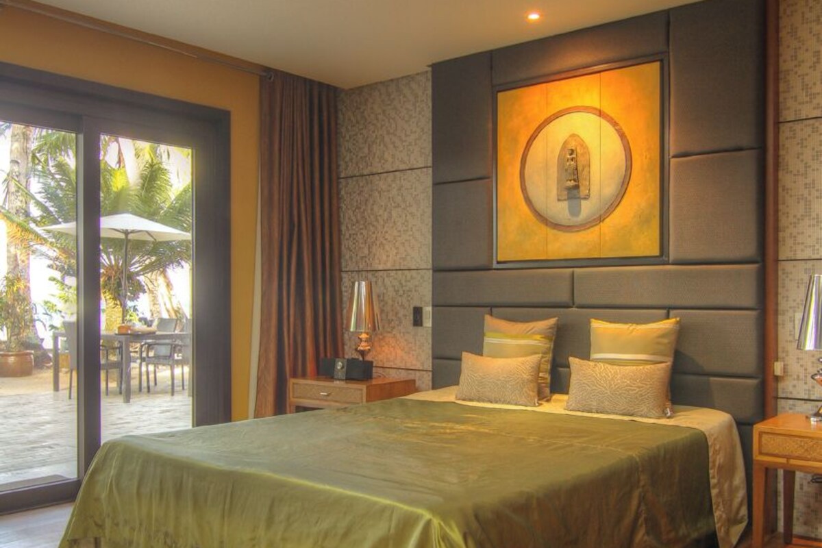 Un dormitorio moderno con una cama grande, una mesita de noche, una lámpara de pared y vistas a un espacio exterior con palmeras.