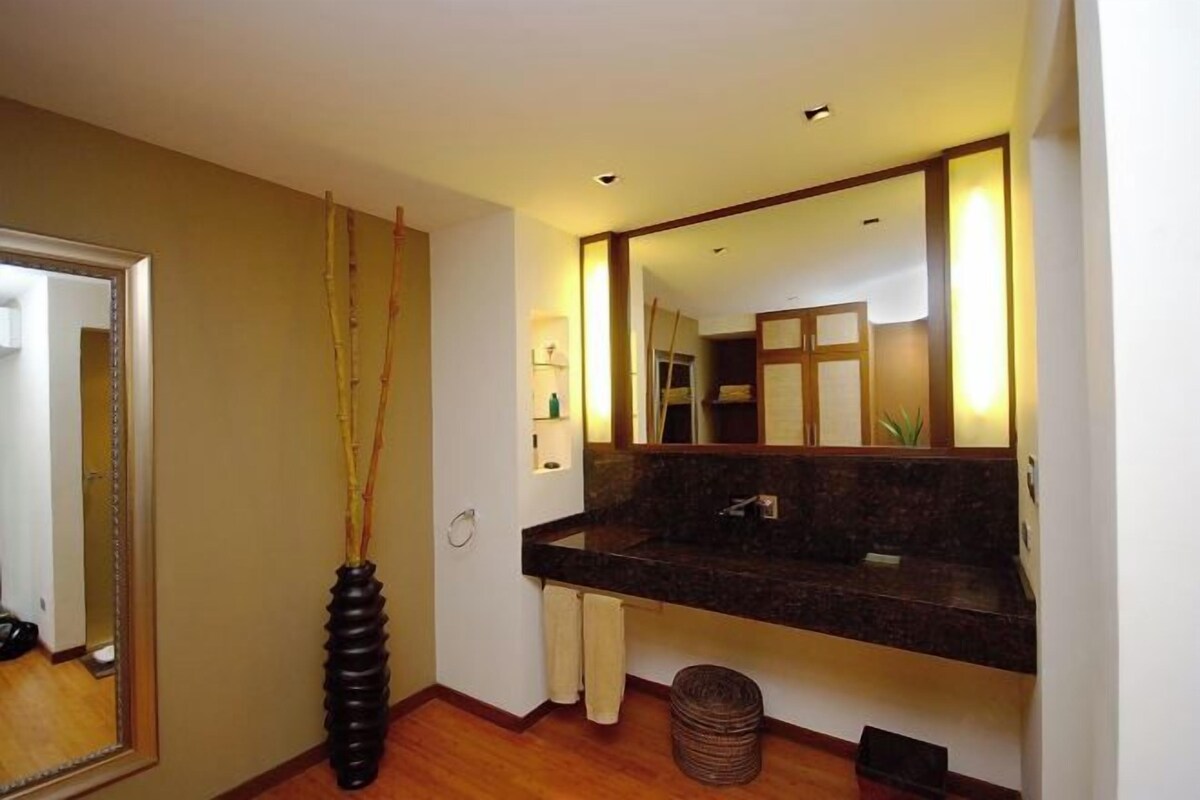 A modern bathroom with a dark granite sink, a large mirror, and a tall vase with branches.