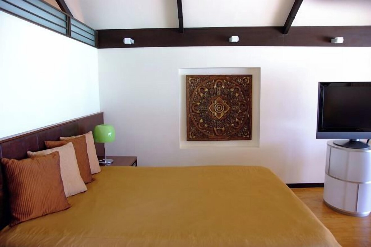 A hotel room with a bed, a television, a decorative wall piece, and a green floor lamp.