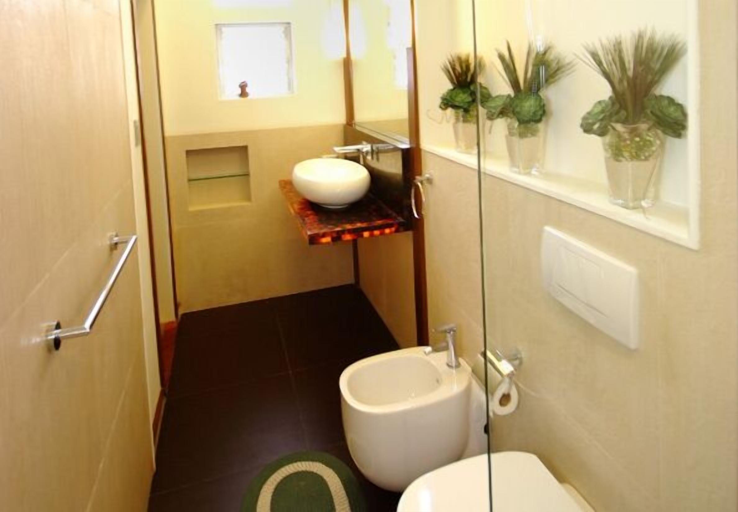 A compact bathroom with a white sink, a bidet, and a toilet. There is a small shelf with a mirror above the sink and a window with plants on the wall.